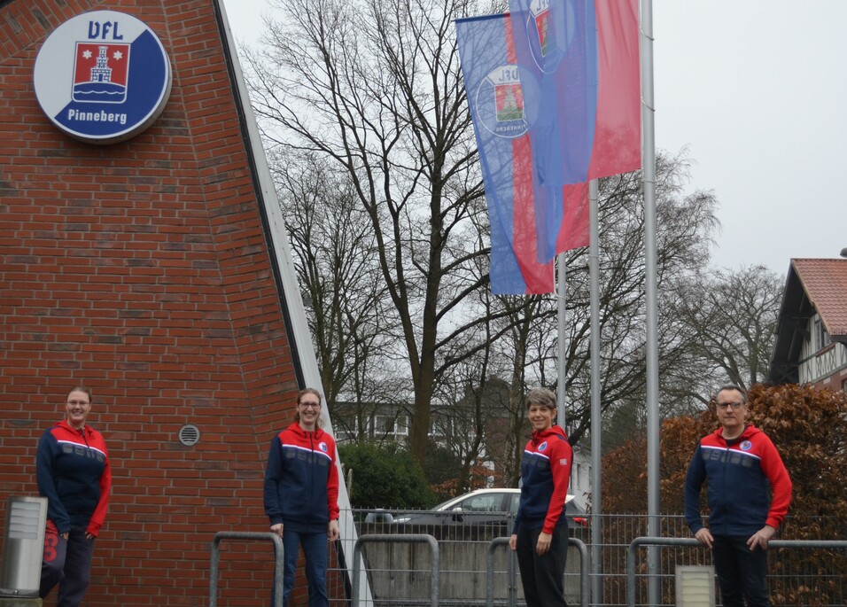 Vier Personen in Sportjacken stehen vor dem DFL Pinneberg-Logo und wehenden Fahnen an einem grauen, bewölkten Tag.