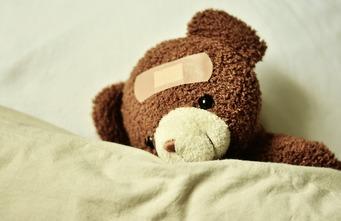 Stoffbär mit Pflaster auf der Stirn liegt auf einem Bett unter einer Decke.