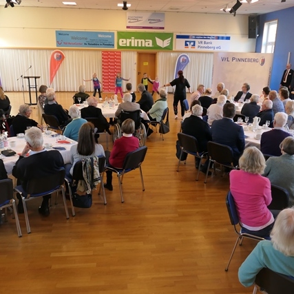 Veranstaltungssaal mit älteren Menschen, die an Tischen sitzen, während Kinder eine Aufführung auf der Bühne zeigen.