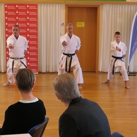 Karate-Demonstration mit fünf Kämpfern in weißen Anzügen auf einer Bühne vor Publikum.