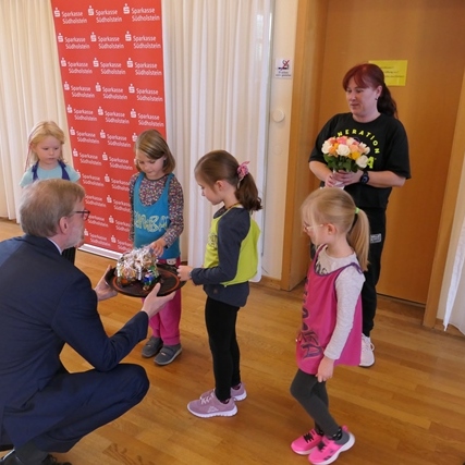 Ein Mann kniet vor vier Kindern und erhält einen bunten Kuchen, während Frauen mit Blumen im Hintergrund stehen.