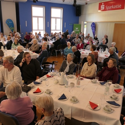 Eine große Gruppe älterer Menschen sitzt an Tischen in einem festlich geschmückten Saal.