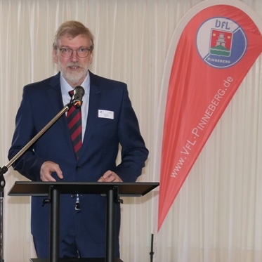 Mann in Anzug spricht am Mikrofon vor einem Tisch, im Hintergrund ein Banner des VfL Pinneberg.