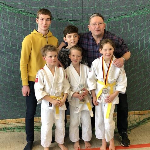 Gruppe von fünf Personen, darunter Kinder in Judoanzügen mit Medaillen, vor einem grünen Netz im Hintergrund.