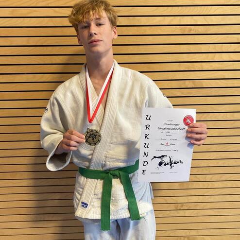 Jugendlicher im weißen Judoanzug mit grüner Gürtelfarbe, hält Medaille und Urkunde für den 1. Platz in der Hamburger Einzelmeisterschaft.
