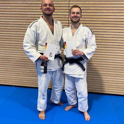 Zwei Judoka in Trainingsanzügen halten Urkunden und stehen auf einer blauen Judomatte vor einer Holzwand.
