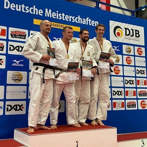 Vier Judoka in weißen Anzügen stehen auf einem Siegertreppchen mit Medaillen und Urkunden vor einer blauen Wand.