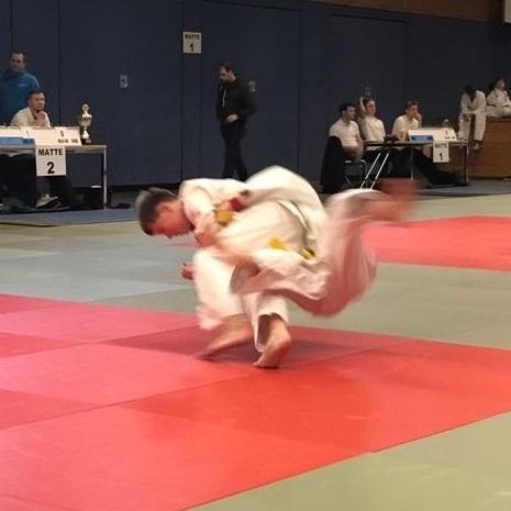 Ein Wettkämpfer im Judogi führt einen Wurf auf der roten Judomatte in einer Turnhalle aus.