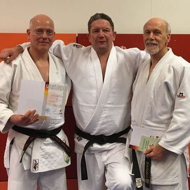 Drei Männer in Judokis, zwei mit Urkunden, stehen vor einer orangefarbenen Wand in einem Trainingsraum.