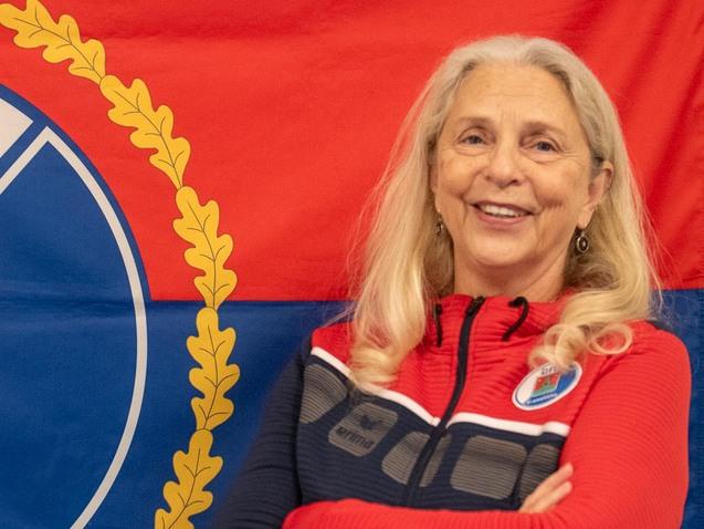 Ältere Frau mit langen, hellen Haaren lächelt vor einer rot-blauen Flagge des VfL Pinneberg.