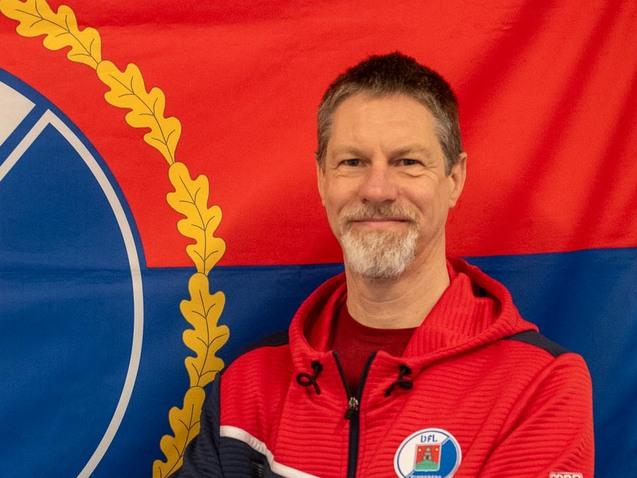Mann mit verschränkten Armen vor einer rot-blauen Flagge mit dem DFL-Logo und dem Wappen von Pinneberg.