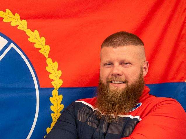 Mann mit Bart steht vor einer rot-blauen Flagge des DFL Pinneberg und lächelt, die Arme verschränkt.