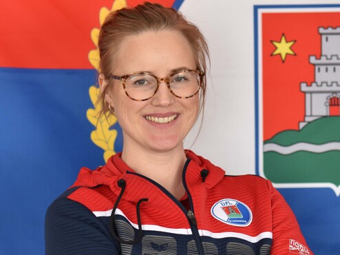 Eine lächelnde Frau mit Brille trägt einen rot-blauen Sportpullover vor einer Fahne im Hintergrund.