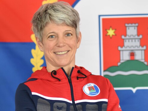 Porträt einer lächelnden Frau mit kurzem, grauem Haar in einem sportlichen Pulli vor einer Flagge.