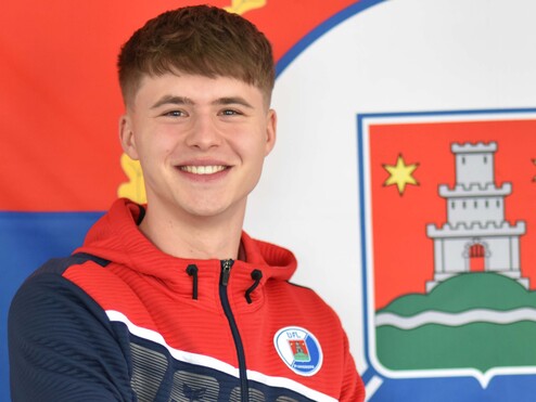Junger Mann in roter und blauer Jacke lächelt vor einer farbigen Flagge mit städtischem Wappen im Hintergrund.