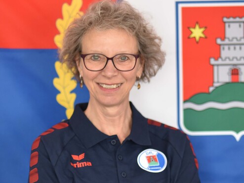 Portrait einer lächelnden Frau mit Brille in einem blauen Poloshirt vor einer Flagge und Wappen im Hintergrund.