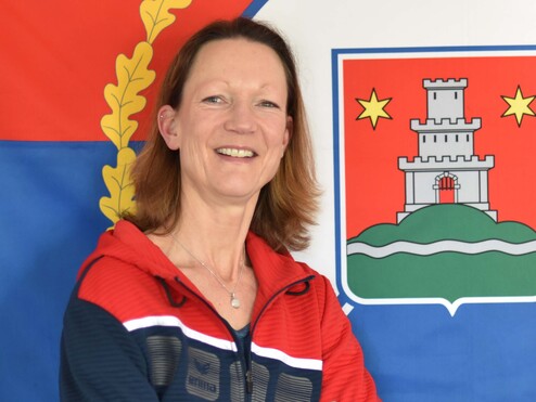 Frau mit sportlichem Outfit lächelt vor einer Flagge und einem Wappen, die für eine Stadt stehen.