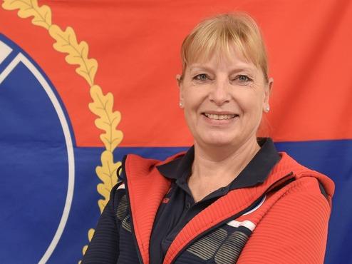 Frau mit blonden Haaren lächelt vor einer Flagge von Pinneberg mit Wappen und roten sowie blauen Elementen.