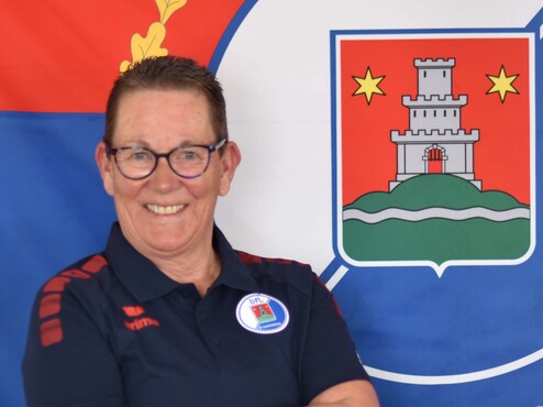 Lächelnde Person mit Brille in Sportkleidung vor einer großen Fahne mit Wappen im Hintergrund.