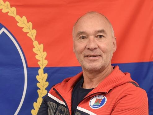 Mann in Sportjacke steht vor einer Flagge mit dem Wappen von Pinneberg und lächelt freundlich in die Kamera.