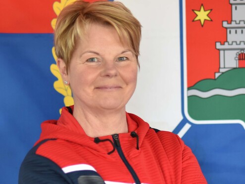 Porträt einer Person mit kurzem, blonden Haar, lächelnd vor rot-blauem Hintergrund mit Wappen. Arme verschränkt.