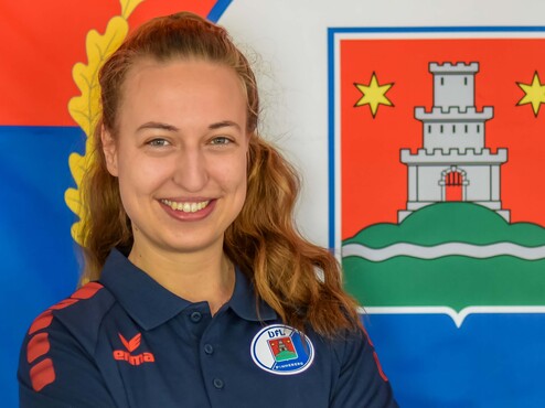 Lächelnde Frau mit gefalteten Armen vor einer farbigen Flagge und Wappen im Hintergrund. Sie trägt ein dunkles Polohemd.