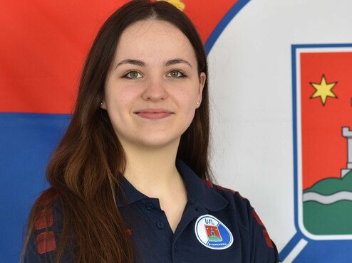 Junge Frau mit langen Haaren, in uniformierter Sportkleidung, lächelt vor einer blauen Flagge mit einem Wappen.