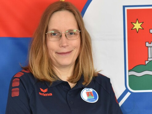 Frau mit Brille in Sportbekleidung, lächelt vor einer Wand mit Stadtwappen und farbigen Flaggen.