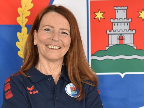 Lächelnde Frau mit langen, roten Haaren, gekreuzten Armen, vor einem bunten Hintergrund mit Wappen und Flagge.