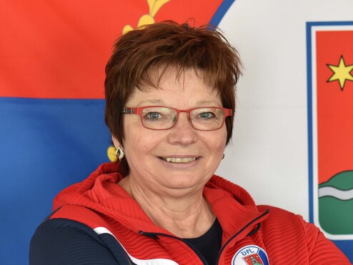 Eine lächelnde Frau mit Brille in einem roten und blauen Sportjacke posiert vor einem Banner in den Farben Blau und Rot.