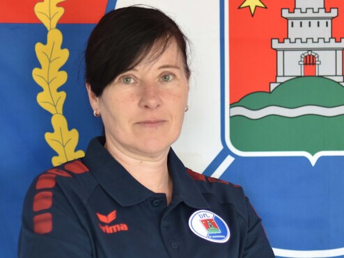 Porträt einer Frau mit verschränkten Armen, im blauen Shirt vor einer Flagge mit Wappen im Hintergrund.