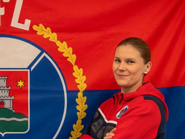 Frau in Sportjacke steht vor einem VFL Pinneberg-Wappen auf rotem und blauem Hintergrund.