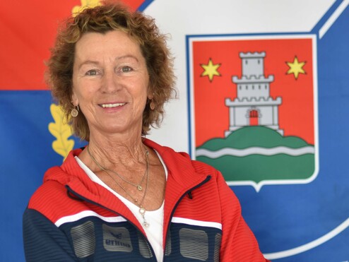 Porträt einer lächelnden Frau in Sportbekleidung vor einer farbigen Flagge mit Wappen im Hintergrund.