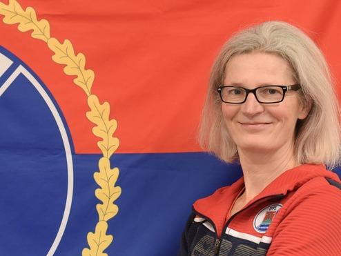 Eine Person mit Brille steht vor einer roten und blauen Flagge mit dem Wappen von Pinneberg.