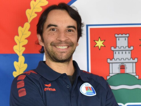 Portrait eines Mannes mit verschränkten Armen, der in einem blauen Poloshirt vor einer Flagge steht.