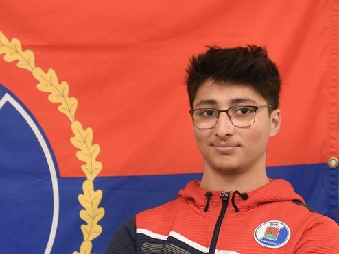 Jugendlicher im roten Sportanzug steht vor einer Wand mit der Flagge eines Sportvereins.