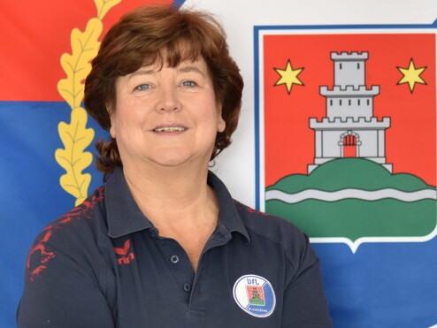 Porträt einer lächelnden Frau mit kurzen braunen Haaren, im Sporthemd vor einer Fahne mit Wappen.