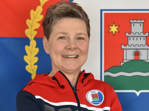 Lächelnde Person mit gekreuzten Armen in einer roten und blauen Jacke vor einem farbenfrohen Hintergrund.