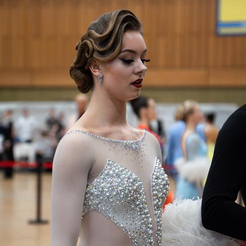 Tanzende Frau in glitzerndem Kleid mit Federn, auf einem Wettkampf im Hintergrund unscharfe Tänzer.