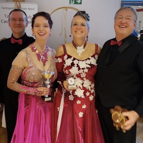 Vierter Tanzsport-Club mit Pokalen, zwei Frauen in glitzernden Kleidern, Männer in Anzügen, fröhliche Stimmung.