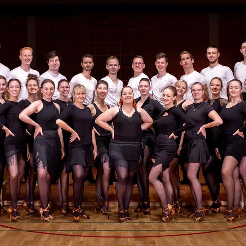 Gruppe von Tänzern in schwarz und weißen Oberteilen, posierend auf einem Parkettboden in einem Sportraum.