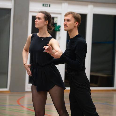 Tanzpaar in eleganter Haltung, beim Training in einer Turnhalle, die Dame trägt ein schwarzes Kleid, der Herr schwarze Kleidung.