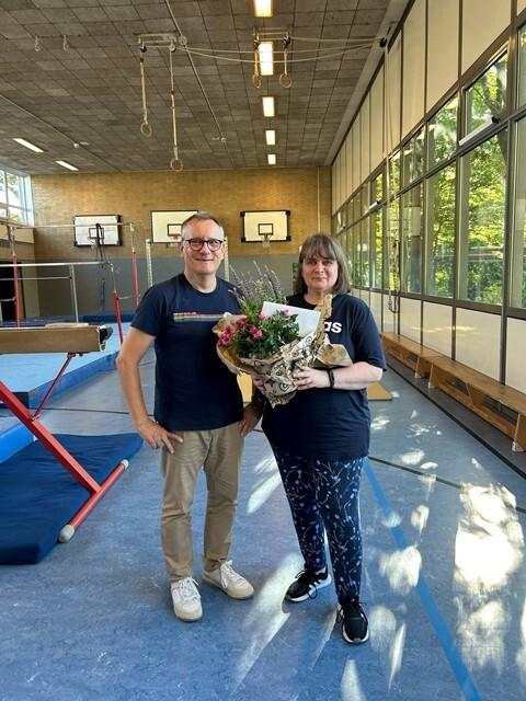 Zwei Personen stehen in einer Turnhalle und halten gemeinsam einen Blumenstrauß. Im Hintergrund sind Turngeräte sichtbar.