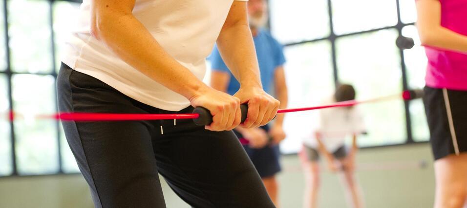 Nahaufnahme von Händen, die einen Widerstandsband in einem Fitnessraum halten, mit Personen im Hintergrund.