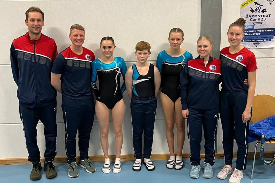 Gruppe von Gymnastikern und Trainern in Sportkleidung, l&auml;chelnd vor einer Wand mit Wettkampfinformationen.