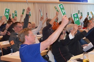 Versammlungsteilnehmer heben gr&uuml;ne &bdquo;Ja&ldquo;-Stimmkarten in die H&ouml;he, w&auml;hrend sie an langen Tischen sitzen.
