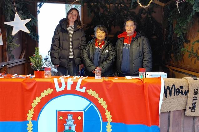 Drei Personen stehen hinter einem Tisch mit einer roten DFL-Flagge und weihnachtlicher Dekoration in einem Weihnachtsstand.