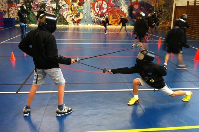 Zwei Kinder im Fencing-Anzug fencen auf einer Sporthalle mit farbenfrohem Graffiti-Hintergrund und orangefarbenen H&uuml;tchen.