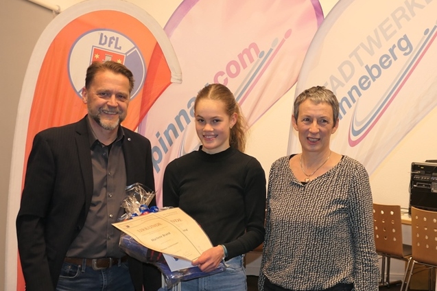 Drei Personen stehen vor bunten Fahnen, eine Jugendliche h&auml;lt Urkunden und einen Blumenstrau&szlig; in den H&auml;nden.