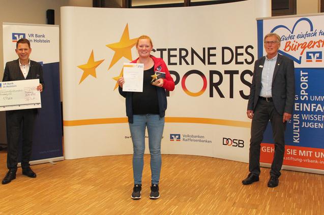 Drei Personen stehen in einem Raum vor einem Banner, eine Person h&auml;lt eine Urkunde und einen Pokal.